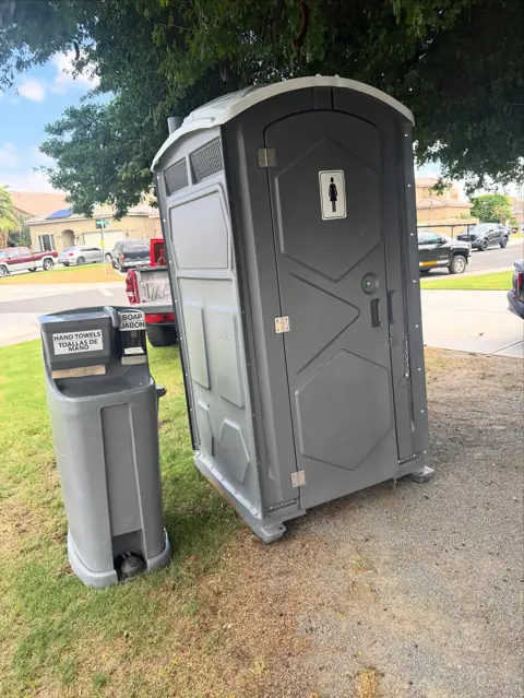 Gray portable toilet next to a foot-pump hand wash station on a lawn